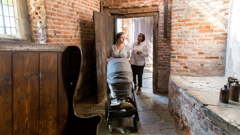 A lady with dark hair and a grey pushchair peers around the corner of a red bricked building, with September sunlight cascading through a window, illuminating the old Brewery, with her retiree mother behind her looking up at the wonky wooden doorframe.